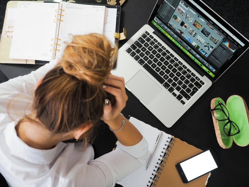 This image shows a picture of a person sitting in front of a desk with a computer, notebook, and phone. They are looking downwards appearing distressed. This image is used from pexels.com for frances may design websites free website audit page.